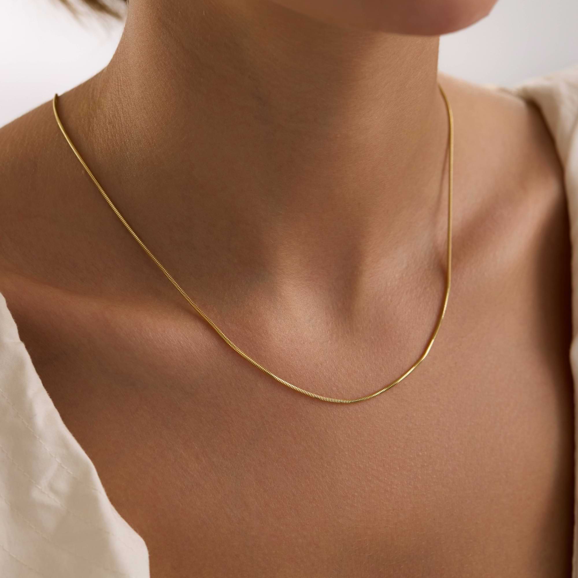 Gold 1.25mm round snake chain necklace on a person's neck with a neutral background
