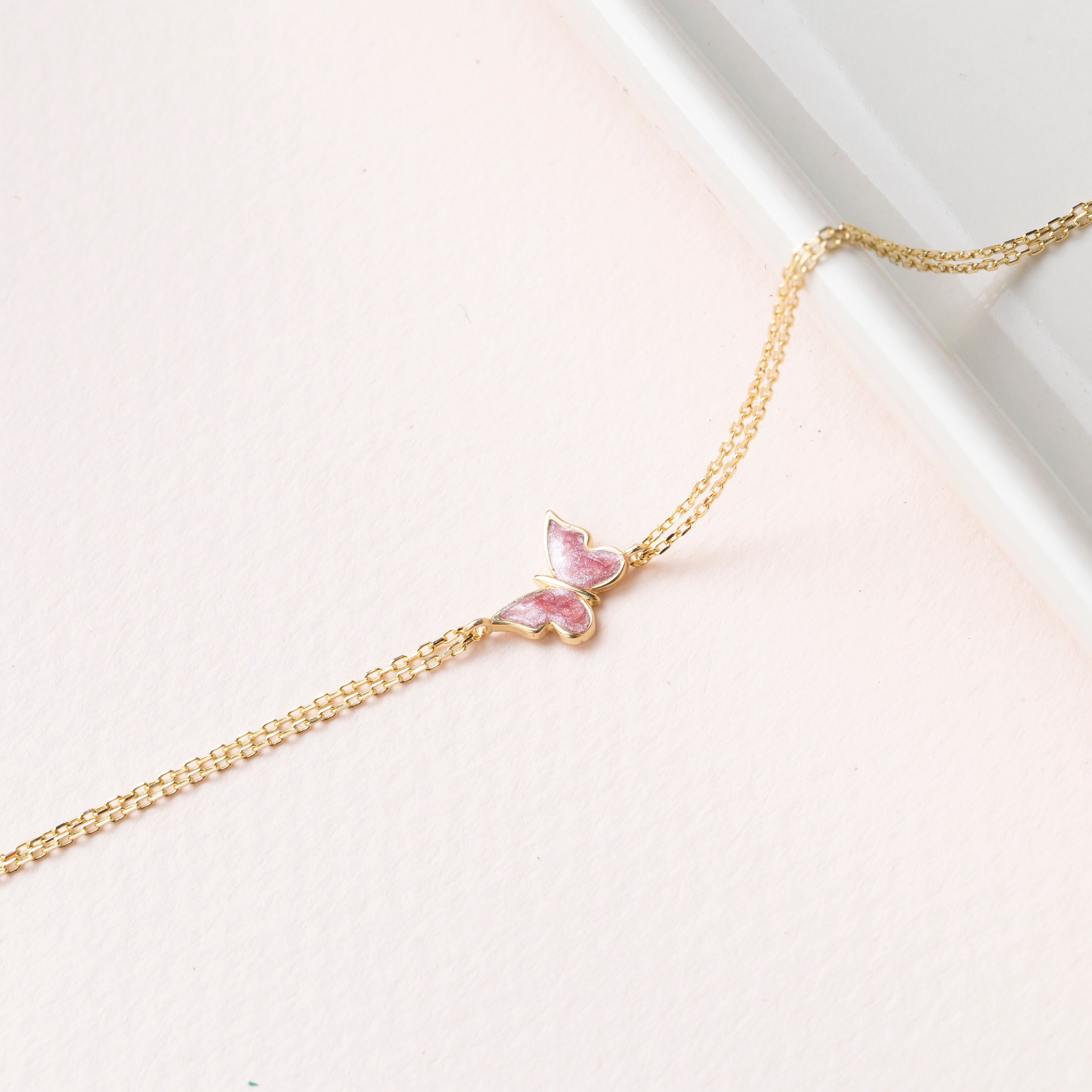 14k gold double chain bracelet with pink enamel butterfly charm, flat lay on blush pink background