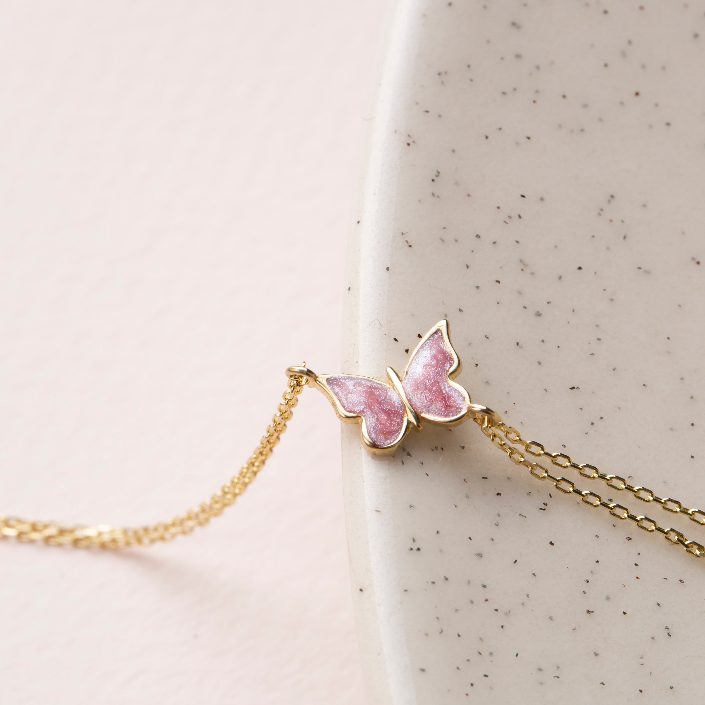 14k gold double chain bracelet with pink enamel butterfly charm displayed on speckled ceramic dish