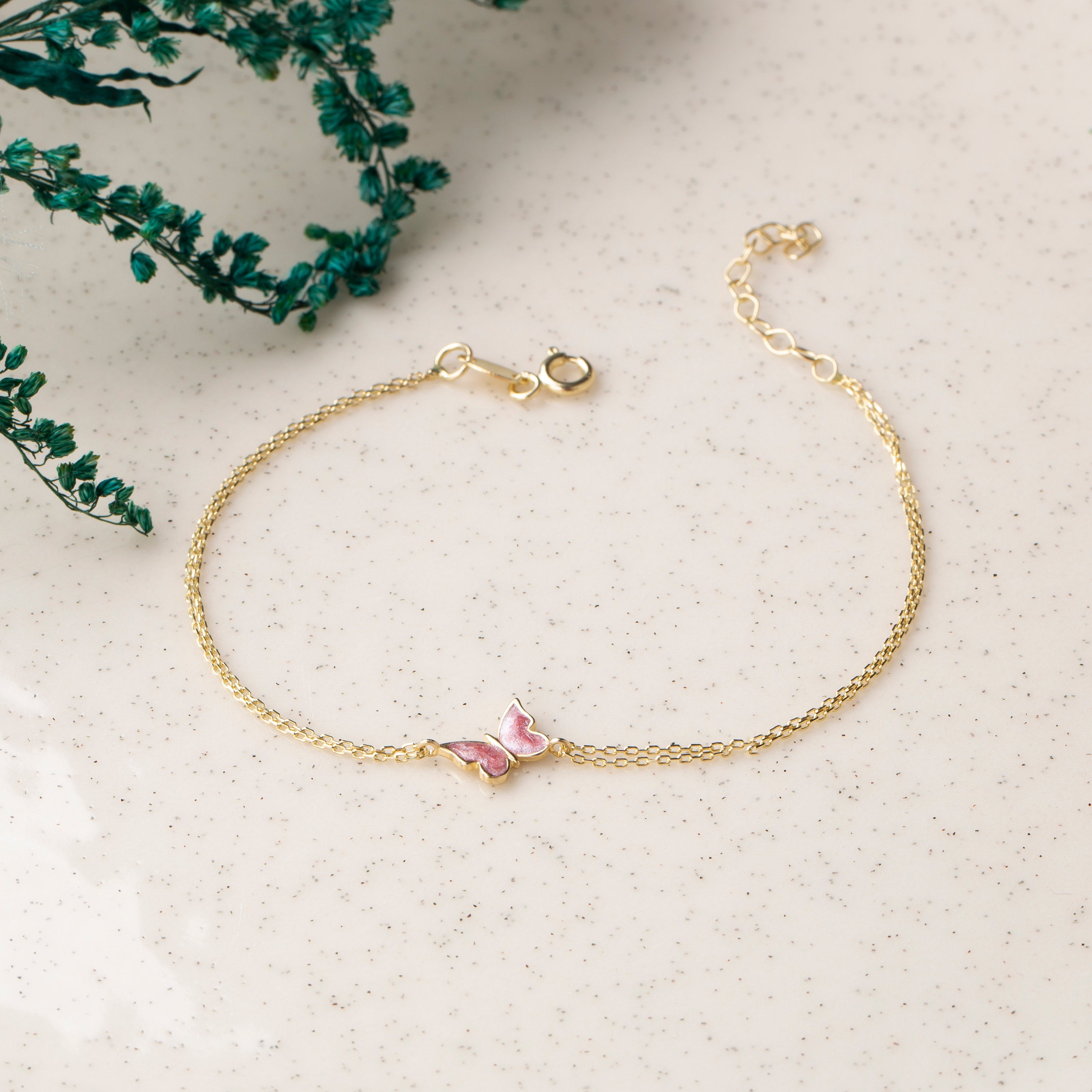 14k gold double chain bracelet with pink enamel butterfly charm and spring ring clasp, styled on speckled stone surface with dried teal flowers