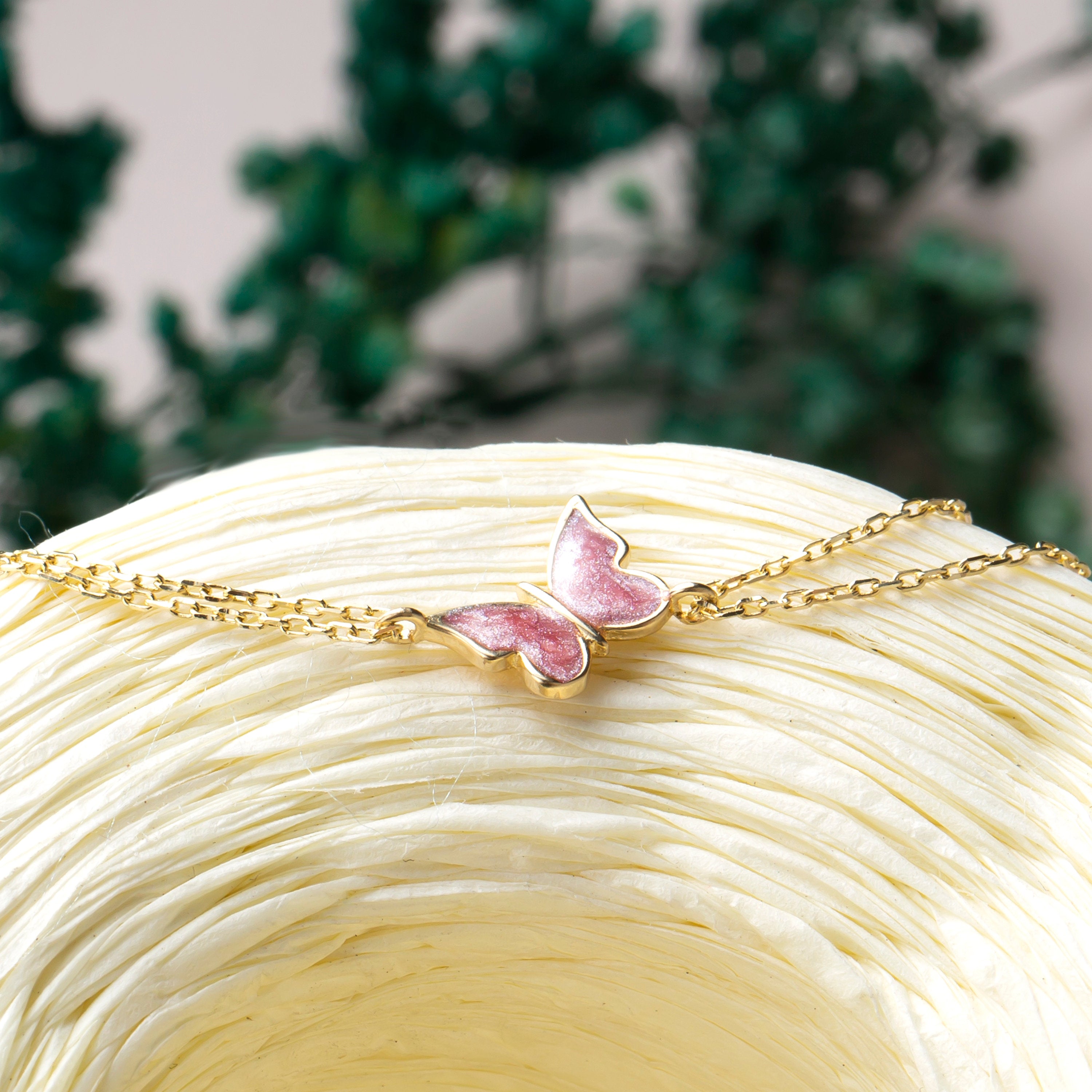 14k Gold bracelet with a pink butterfly charm on a white surface with greenery in the background
