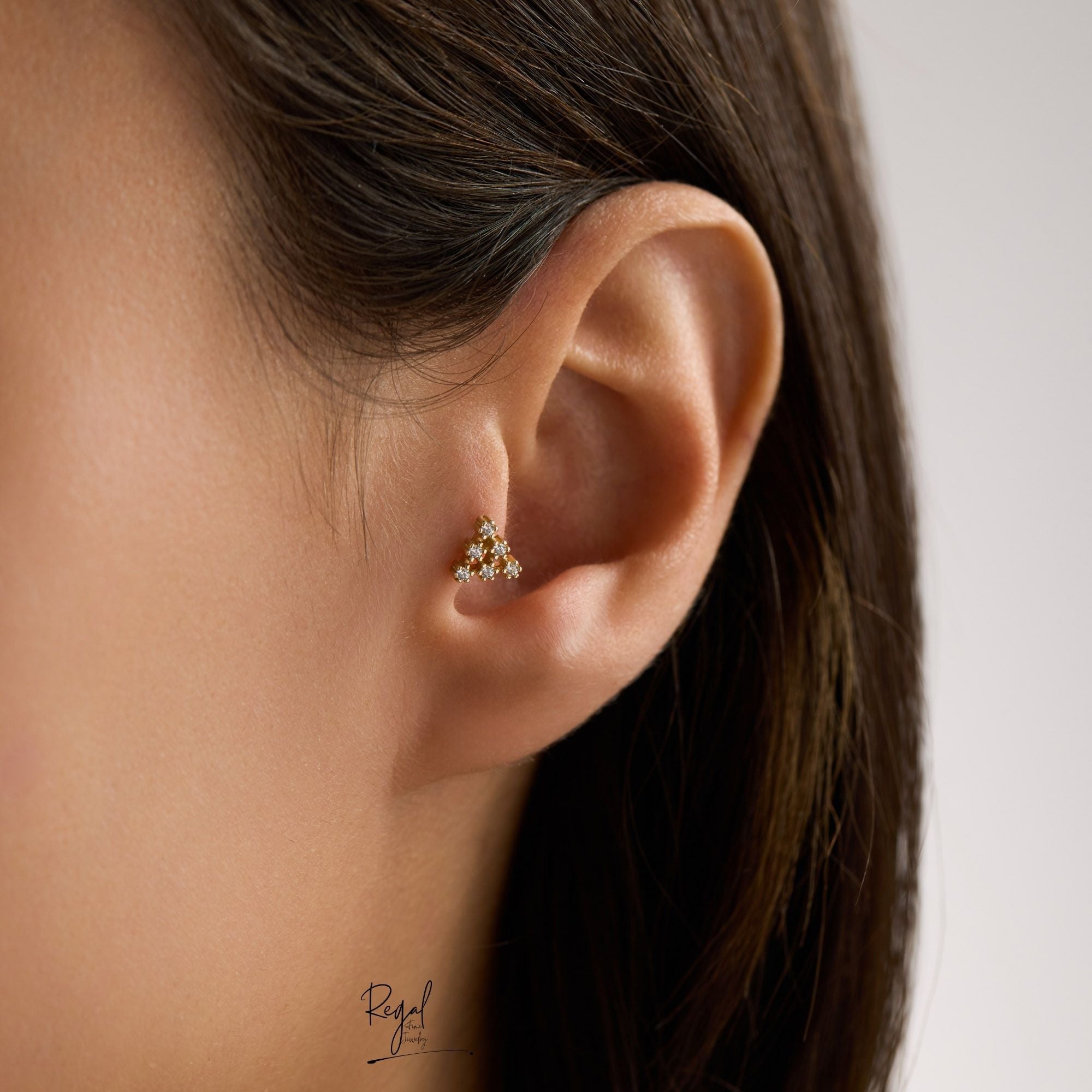 Close-up of an ear tragus wearing a small 14K solid gold triangle CZ cluster tragus piercing with 'Regal Fine Jewelry' branding.