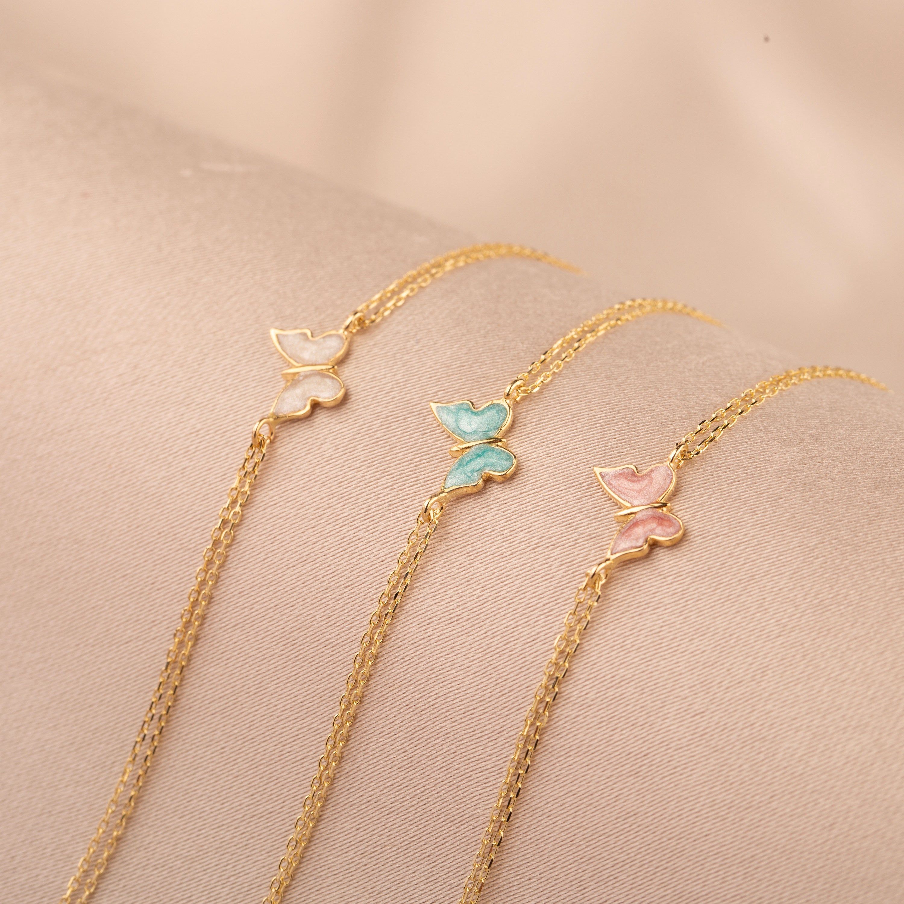 14k gold double chain butterfly bracelet in three enamel color variants — white, turquoise, and pink — displayed on blush fabric
