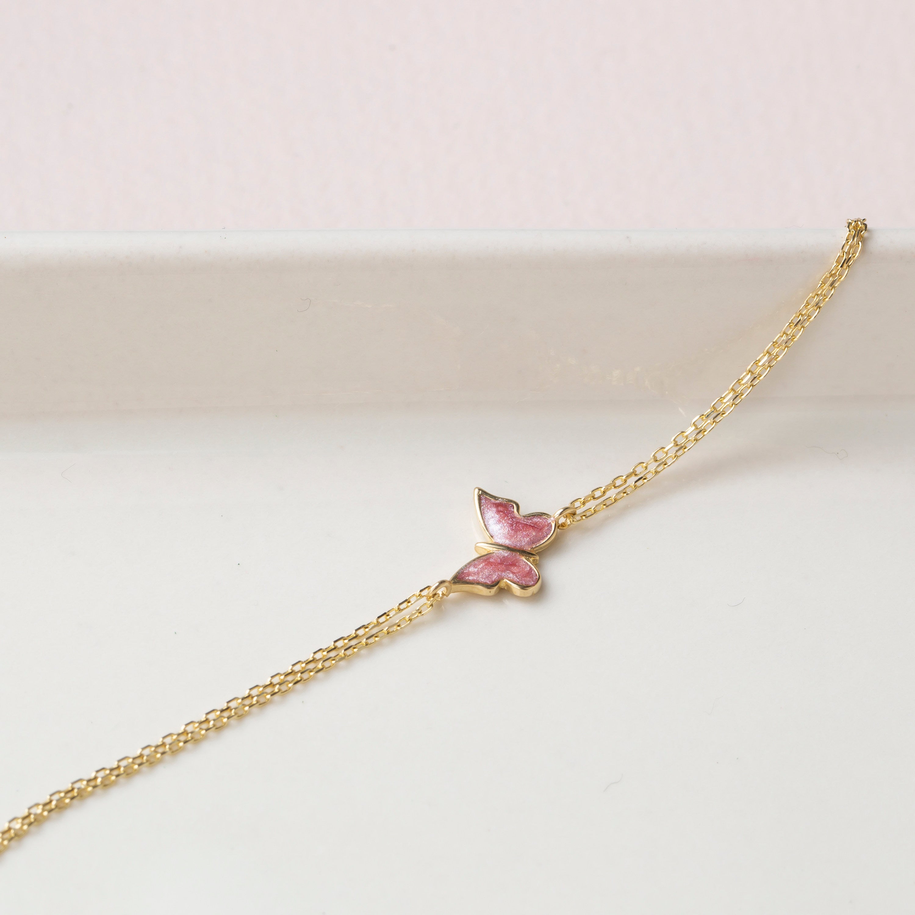 14k gold double chain bracelet with pink enamel butterfly charm, close-up detail on white marble surface