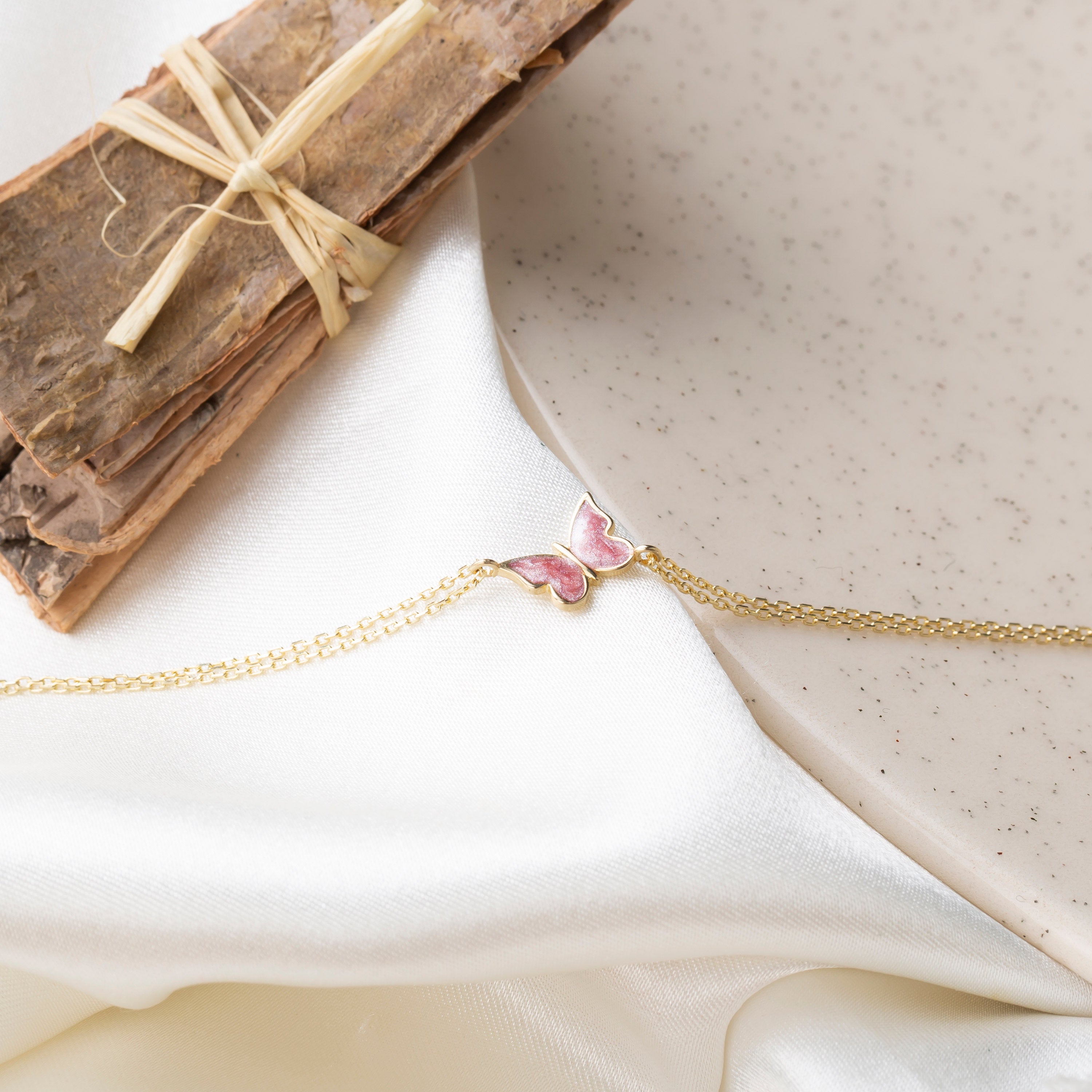 14k gold double chain bracelet with pink enamel butterfly charm styled on white silk fabric with rustic bark bundle